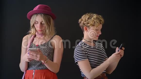 两个年轻漂亮的金发美女朋友在电话里发短信站在黑色背景上一个迷人的女人视频的预览图