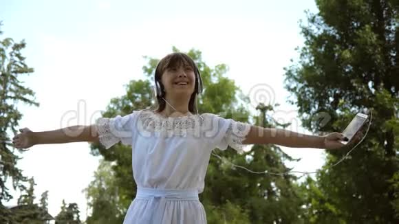 快乐的女孩戴着耳机微笑手里拿着智能手机在夏天的公园里跳舞和旋转慢动作视频的预览图