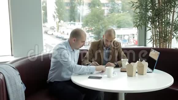 两个年轻人在窗边的一家咖啡馆吃午饭喝咖啡吃面条讨论生意工作笔记本电脑视频的预览图