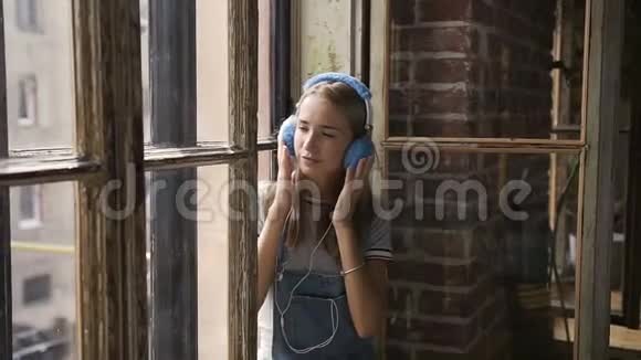 窗户背景上年轻迷人的女孩用耳机听音乐的肖像一个漂亮的女孩坐在附近视频的预览图
