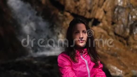 穿着红色夹克的瀑布背景上的年轻美女坐在石头上享受生活的女人视频的预览图