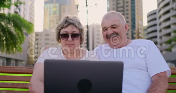 今年夏天一对快乐的老夫妇坐在一个现代城市的长凳上手提电脑在棕榈树上视频的预览图