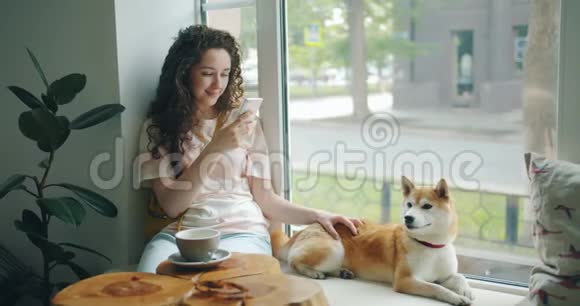 有魅力的女人用智能手机坐在窗台上和小狗在咖啡馆里视频的预览图