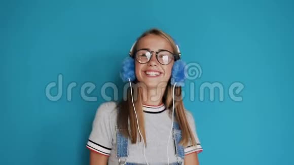 戴眼镜的漂亮女孩正在蓝色的室内跳舞并随着音乐的节奏在耳机里听音乐视频的预览图