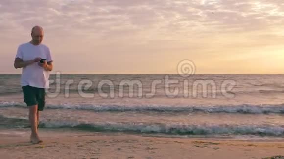 成熟的男人在靠近海洋的沙滩上散步时用电话视频的预览图