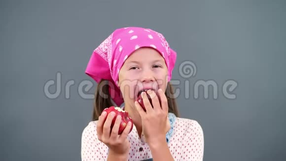 可爱的农民女孩手里拿着两个成熟的大红苹果在工作室里吃咬在灰色背景上视频的预览图