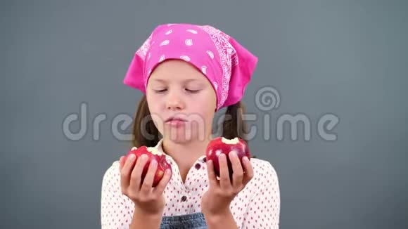 可爱的农民女孩手里拿着两个成熟的大红苹果在工作室里吃咬在灰色背景上视频的预览图