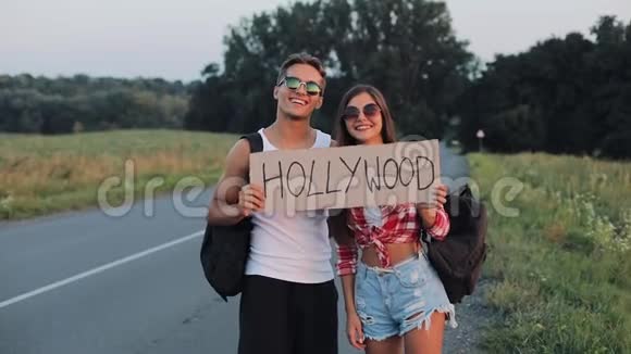 年轻的旅行情侣相爱站在路上举着好莱坞的标志快乐的微笑的朋友们正在注视视频的预览图