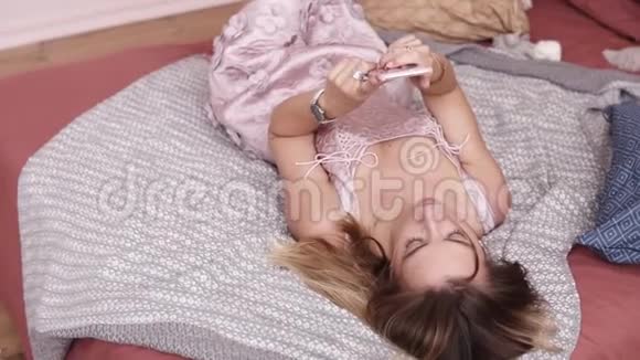 一个白人女孩躺在床上穿着一件非常花哨的衣服在智能手机上打字和滚动一些东西视频的预览图