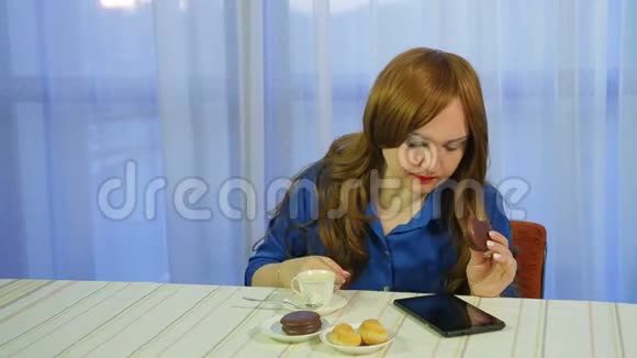 年轻的棕色头发的女人在一张桌子上的咖啡馆里喝着咖啡和甜点读着平板电脑上的新闻视频的预览图