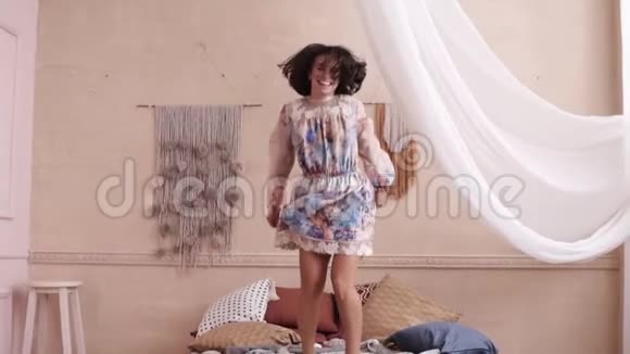 快乐的女人穿着一件可爱的花裙在床上跳跃有白色窗帘的房间前段录像视频的预览图