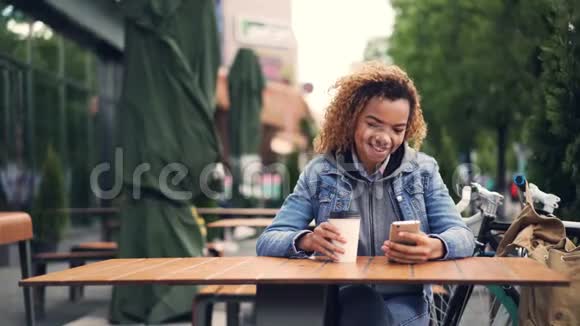 快乐的非裔美国女孩坐在公园的户外咖啡馆里用智能手机触摸屏幕微笑视频的预览图