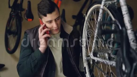 严肃的年轻人自行车修理工正在检查和转动车轮同时在手机上交谈自行车维修视频的预览图
