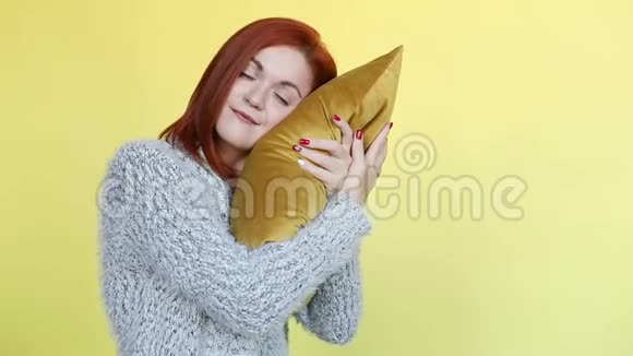 穿着毛衣抱着枕头在黄色背景上摆姿势的红发女人微笑视频的预览图