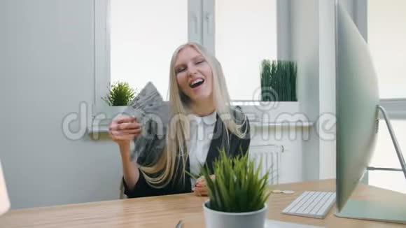 微笑的商业女人拿着一大堆现金美丽快乐的金发女性坐在办公室里用电脑视频的预览图