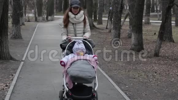 漂亮的女人和她的小女儿一起走在公园里推着婴儿车妈妈带着婴儿车在公园散步视频的预览图