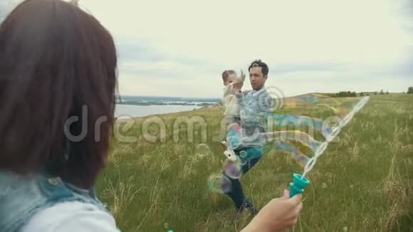 一个年轻的家庭和一个孩子在田里玩耍一个母亲吹泡泡一个父亲带着一个孩子抓住他们笑着视频的预览图