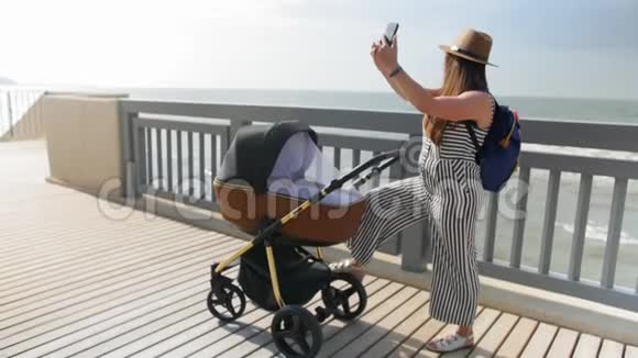 一位年轻的母亲带着婴儿车沿着木板路走用手机和自拍全垒打视频的预览图