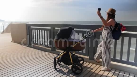 一位年轻的母亲带着婴儿车沿着木板路走用手机和自拍全垒打视频的预览图