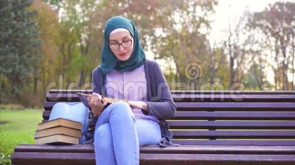 可爱的穆斯林女孩戴着头巾在日落时坐在长凳上看书视频的预览图