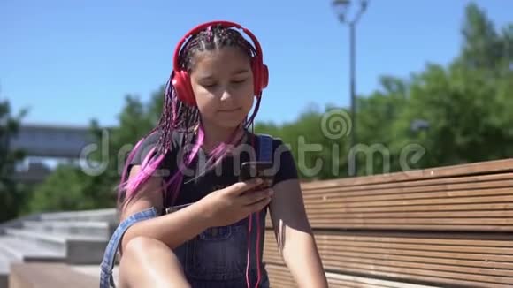 女孩听音乐耳机使用智能手机坐在长凳上心情愉快的夏季公园慢动作视频的预览图