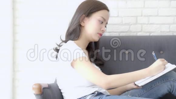 美丽的亚洲女人微笑着坐在沙发上学习在家学习写笔记本视频的预览图