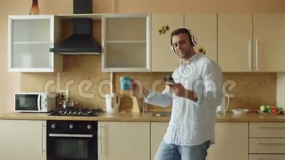 一个英俊的年轻人戴着耳机在家里的厨房里跳舞唱歌在假期里玩得开心视频的预览图