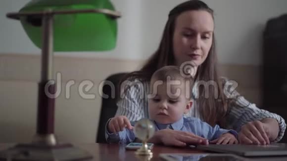 母亲和小儿子坐在室内桌子上的笔记本电脑的肖像忙碌的年轻母亲在家工作这就是视频的预览图