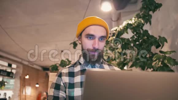 戴着黄色帽子的嬉皮士使用笔记本电脑坐在咖啡馆或同事间工作视频的预览图