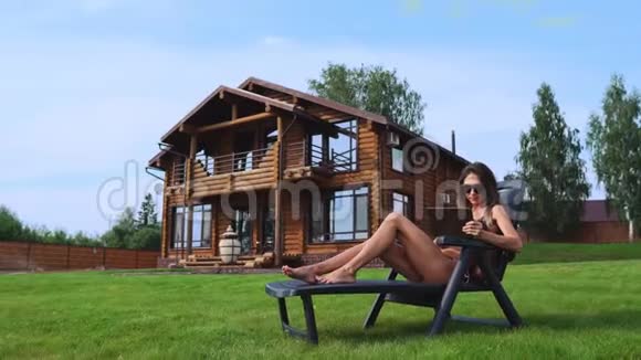 一位美丽的女士躺在她豪宅后院的一个阳光休息室里正在用平板电脑做网上购物视频的预览图