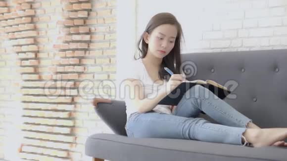 漂亮的亚洲女人坐在沙发上学习在家里写笔记本做家庭作业视频的预览图