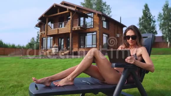 一位身穿泳装的美丽黑发女子躺在她别致的豪宅附近的日光休息室里工作时她微笑着大窗户视频的预览图