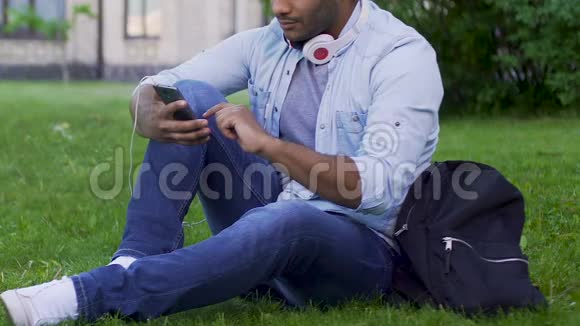 混血小子坐在草地上带着手机和耳机应用程序视频的预览图