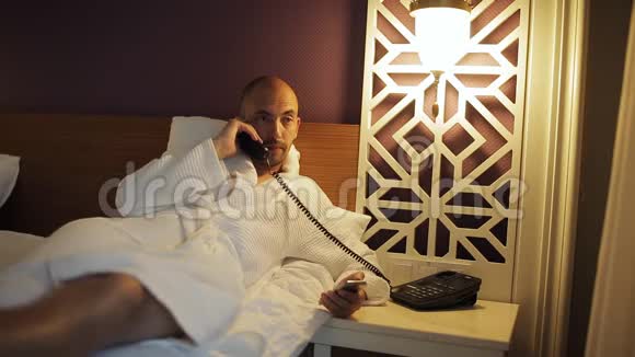 一个穿浴袍的男人躺在床上打电话给酒店的接待处视频的预览图