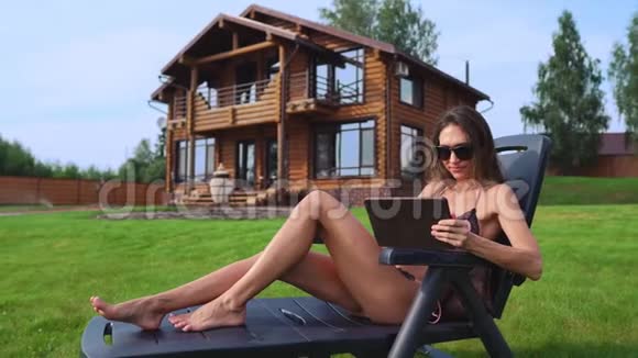 一位身穿泳装身穿长裙坐在她别致的豪宅旁的躺椅上的漂亮黑发女郎一边大摇大摆地微笑着视频的预览图