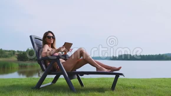 一位在平板电脑上工作的女士度假在湖边的海滩上享受日光浴视频的预览图