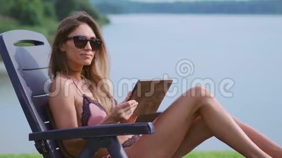 一位在平板电脑上工作的女士度假在湖边的海滩上享受日光浴视频的预览图