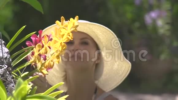 美丽的女人嗅着夏天花园里盛开的花朵的香气戴帽子的女孩享受盛开的兰花的芬芳视频的预览图