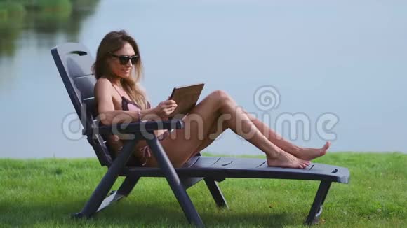穿着泳衣戴着平板电脑的女人躺在阳光休息室里手指触摸着平板电脑的屏幕微笑着视频的预览图