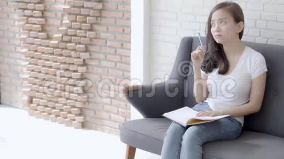 漂亮的亚洲女人坐在沙发上学习在家里写笔记本做家庭作业视频的预览图