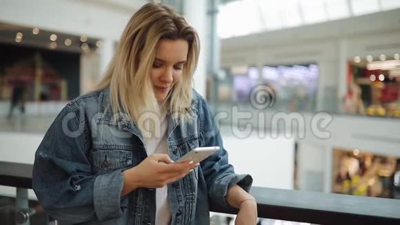 年轻女子微笑着在她的智能手机里键入一些东西站在商场里拿着袋子视频的预览图