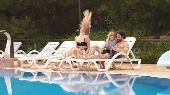 婴儿和父母在度假村游泳池附近的日光浴中放松时尚女孩情侣度假村家庭视频的预览图