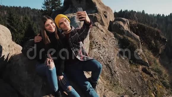 恋爱中的年轻夫妇坐在岩石上拍一张自拍参观著名旅游景点的男女视频的预览图