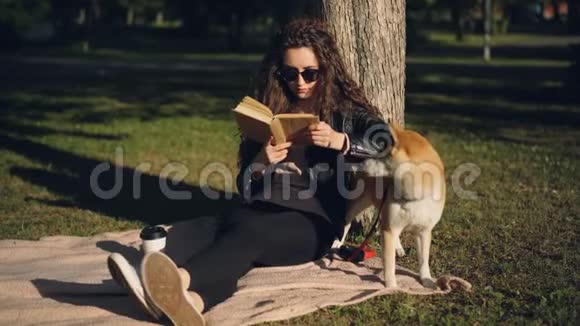 美丽的年轻女子正在看书坐在公园树下的毯子上微笑着而她的家狗却在笑视频的预览图