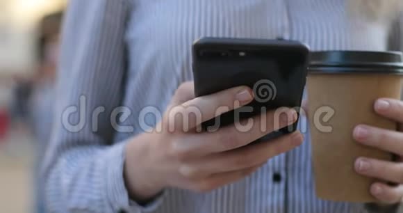 年轻的商务女性穿着衬衫手里拿着手机和咖啡发短信近距离观看视频的预览图