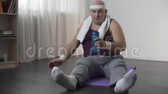 运动服中的胖男人经过艰苦的锻炼后坐在垫子上喝水视频的预览图