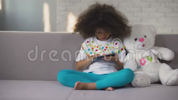 非裔美国女孩坐在沙发上拿着最喜欢的玩具玩手机视频的预览图