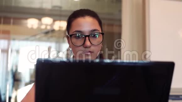戴着眼镜戴着深色头发用电脑和手机跑步的女商人坐在办公室的桌子上表现得很好视频的预览图