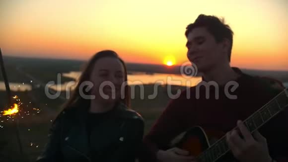 年轻快乐的一对情侣在日落时分在户外用吉他弹唱歌曲视频的预览图
