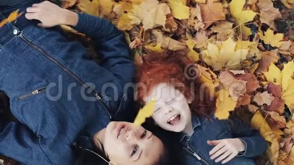 一位美丽的母亲和她可爱的女儿躺在秋天公园的一片黄叶上他们看着镜头俯视视频的预览图